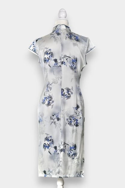 White silk Cheongsam dress featuring a blue and grey ink-wash floral pattern, mandarin collar, and traditional frog buttons on a mannequin.