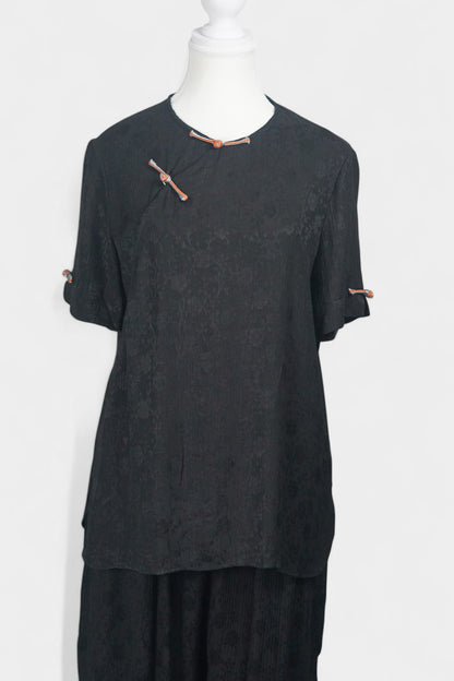 Black textured silk two-piece set featuring a short-sleeve top with contrast frog buttons and matching lantern pants.