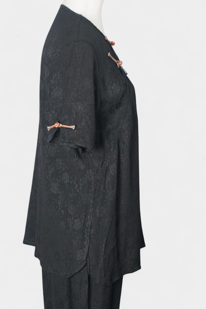 Black textured silk two-piece set featuring a short-sleeve top with contrast frog buttons and matching lantern pants