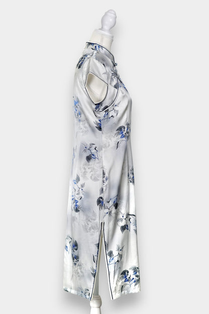 White silk Cheongsam dress featuring a blue and grey ink-wash floral pattern, mandarin collar, and traditional frog buttons on a mannequin.
