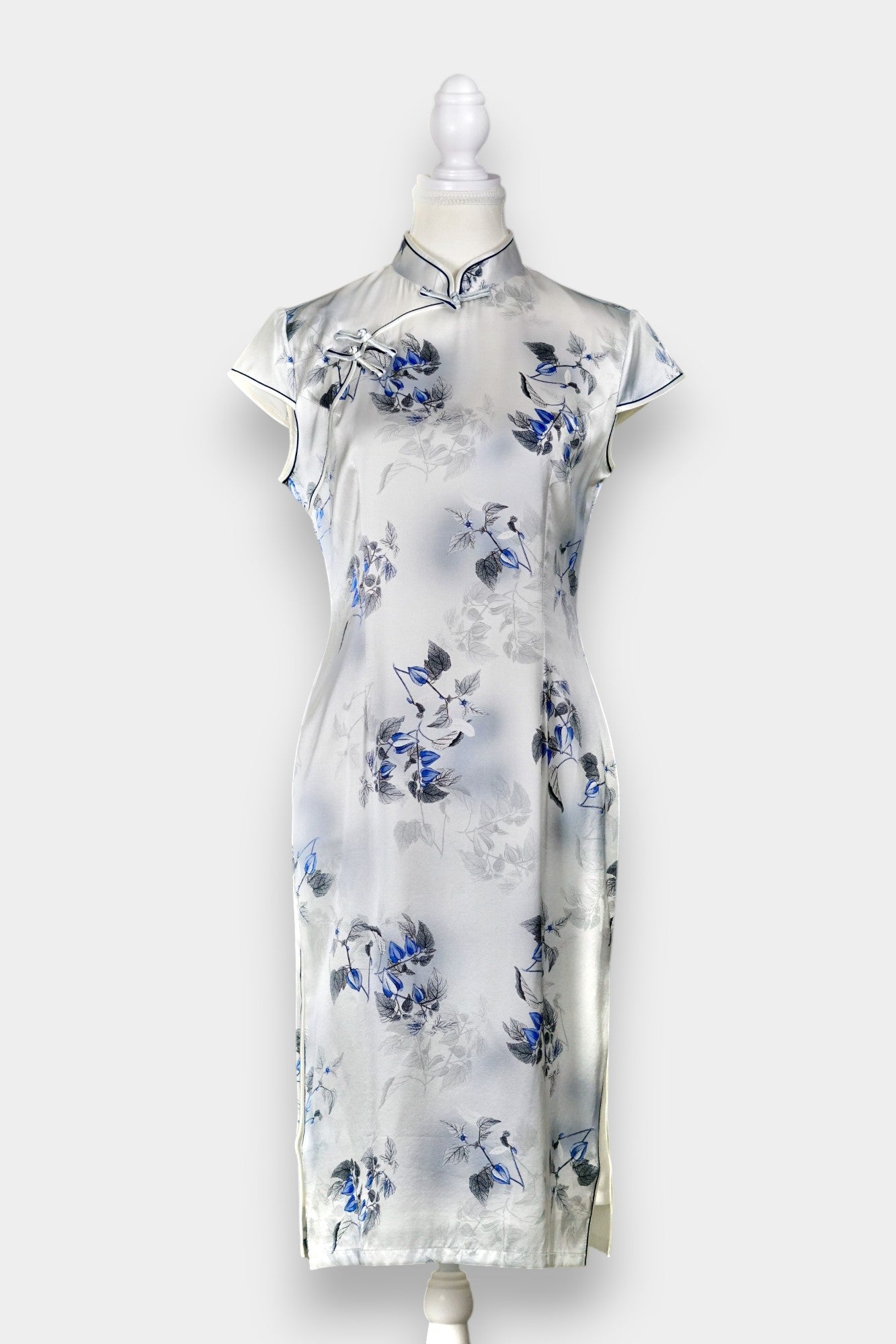 White silk Cheongsam dress featuring a blue and grey ink-wash floral pattern, mandarin collar, and traditional frog buttons on a mannequin.