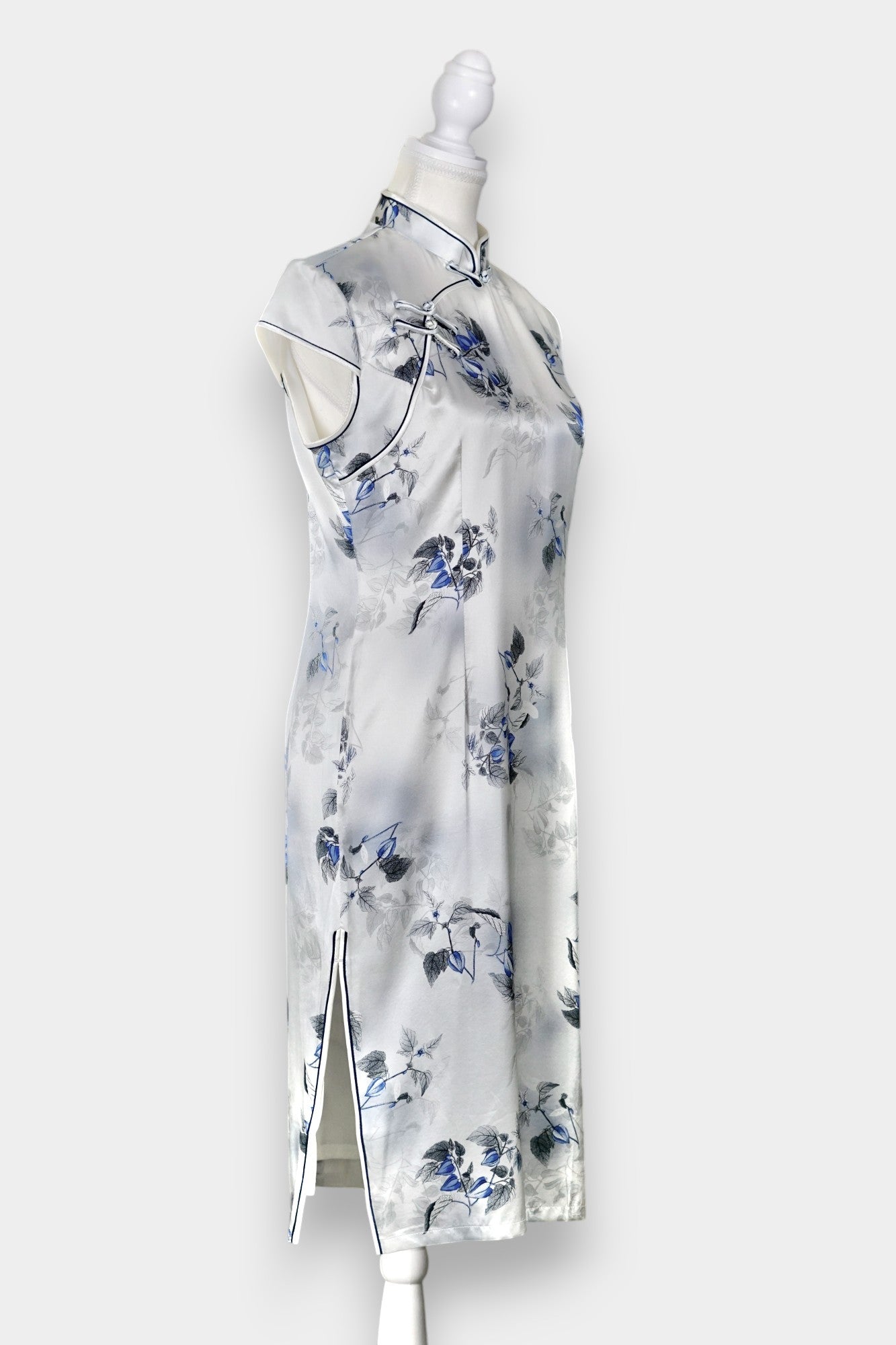 White silk Cheongsam dress featuring a blue and grey ink-wash floral pattern, mandarin collar, and traditional frog buttons on a mannequin.