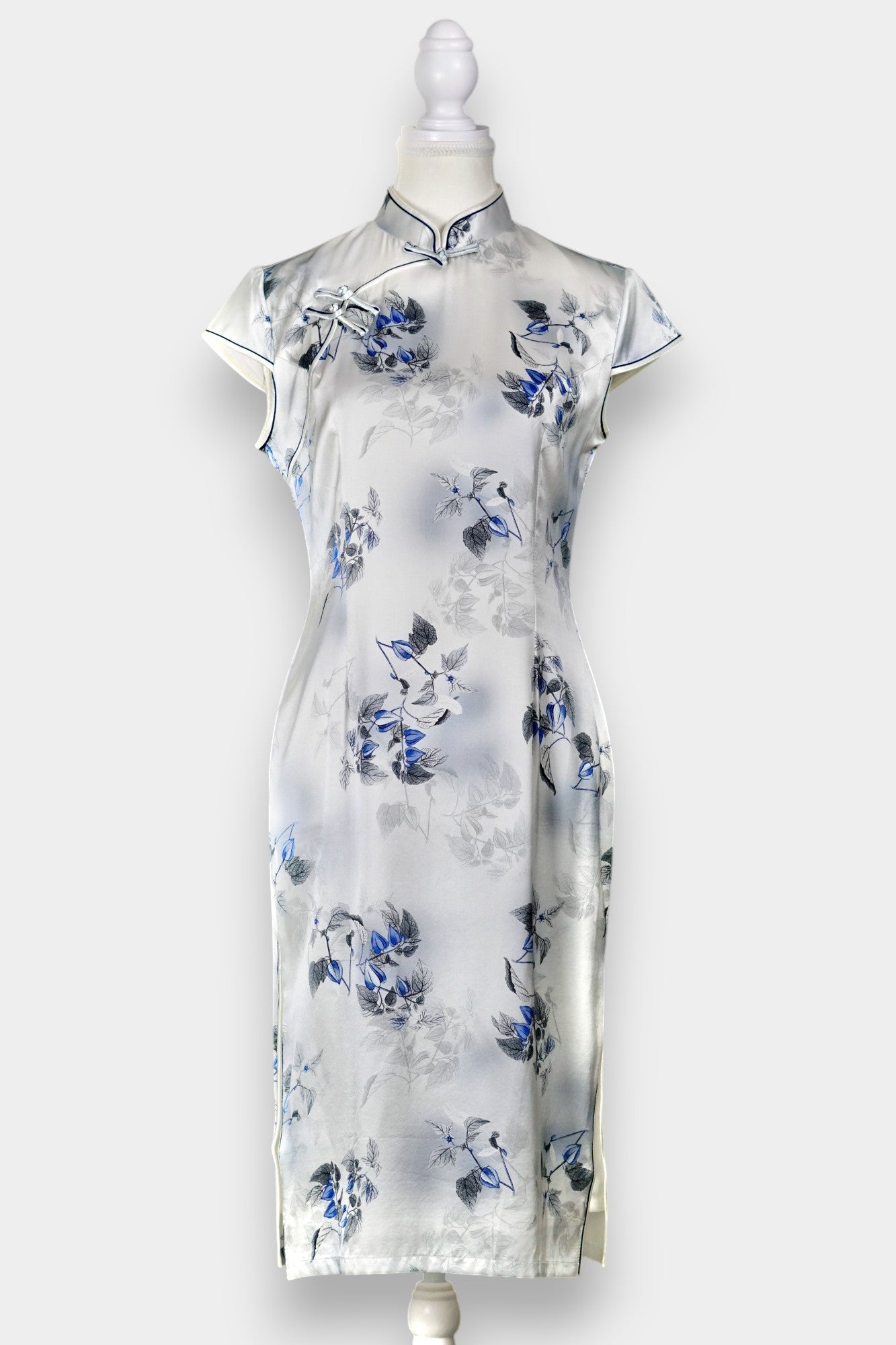 White silk Cheongsam dress featuring a blue and grey ink-wash floral pattern, mandarin collar, and traditional frog buttons on a mannequin.