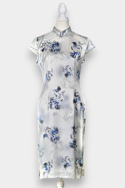 White silk Cheongsam dress featuring a blue and grey ink-wash floral pattern, mandarin collar, and traditional frog buttons on a mannequin.