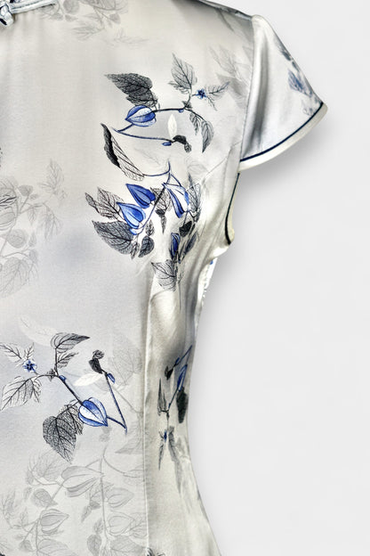 White silk Cheongsam dress featuring a blue and grey ink-wash floral pattern, mandarin collar, and traditional frog buttons on a mannequin.