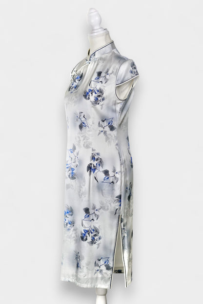 White silk Cheongsam dress featuring a blue and grey ink-wash floral pattern, mandarin collar, and traditional frog buttons on a mannequin.