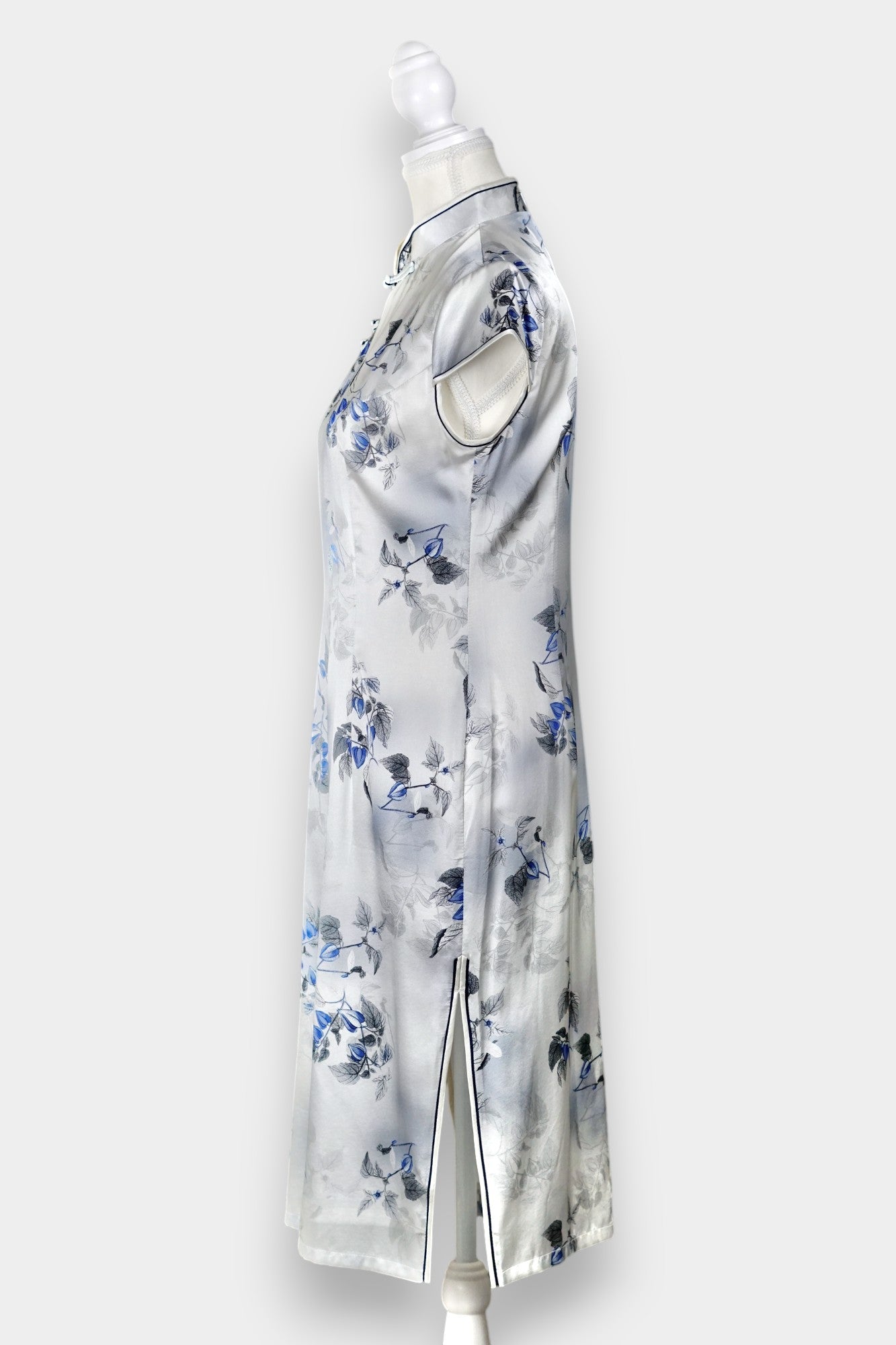 White silk Cheongsam dress featuring a blue and grey ink-wash floral pattern, mandarin collar, and traditional frog buttons on a mannequin.