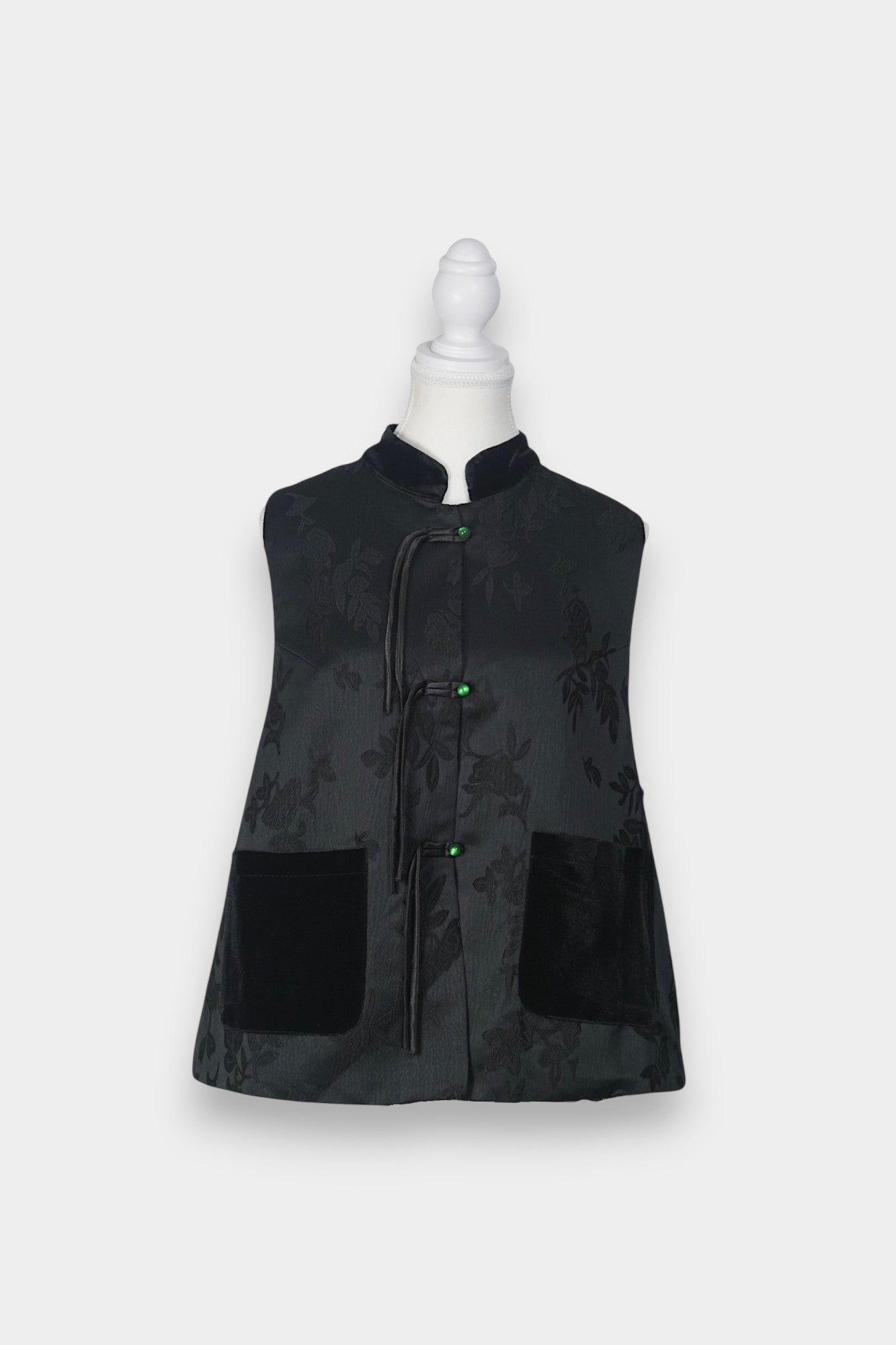 Sleeveless black vest featuring subtle floral jacquard patterns, a velvet Mandarin collar, two velvet patch pockets, and three decorative frog buttons with green beads and long tassels.