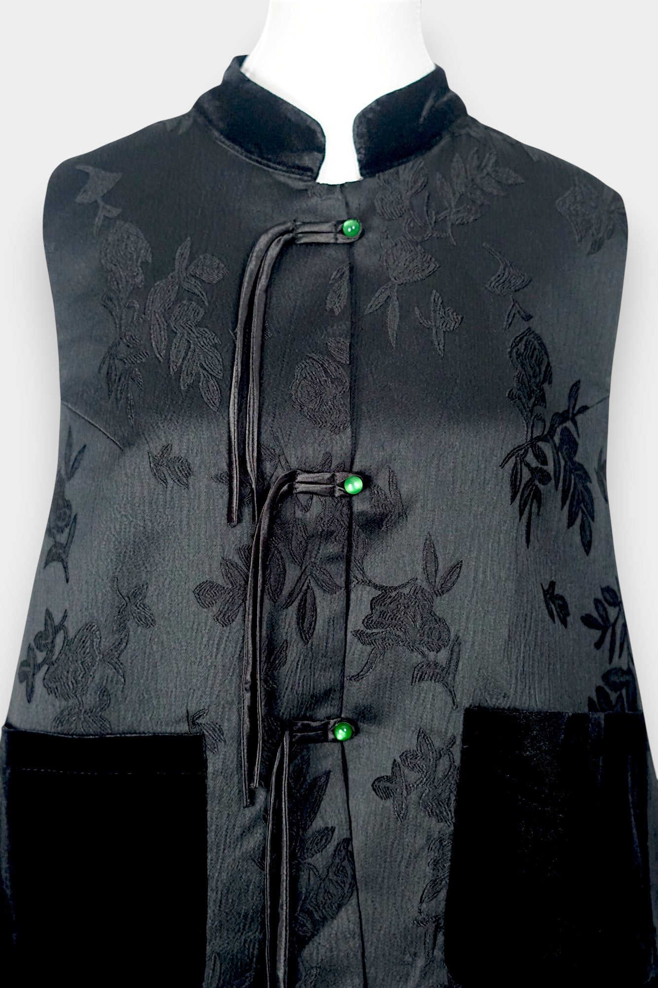 Sleeveless black vest featuring subtle floral jacquard patterns, a velvet Mandarin collar, two velvet patch pockets, and three decorative frog buttons with green beads and long tassels.