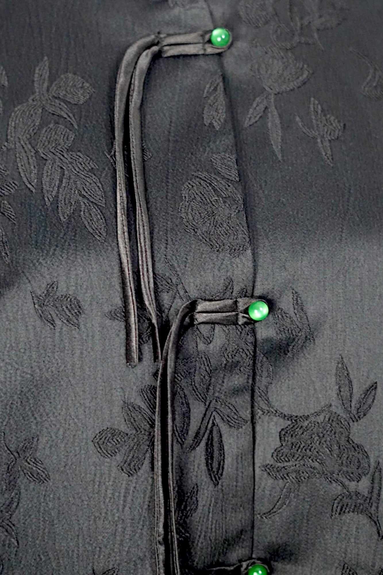 Sleeveless black vest featuring subtle floral jacquard patterns, a velvet Mandarin collar, two velvet patch pockets, and three decorative frog buttons with green beads and long tassels.