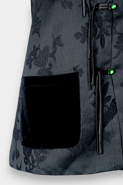 Sleeveless black vest featuring subtle floral jacquard patterns, a velvet Mandarin collar, two velvet patch pockets, and three decorative frog buttons with green beads and long tassels.